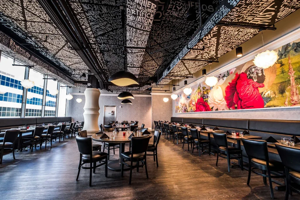 Modern restaurant interior with colorful mural, unique ceiling text, and set tables.