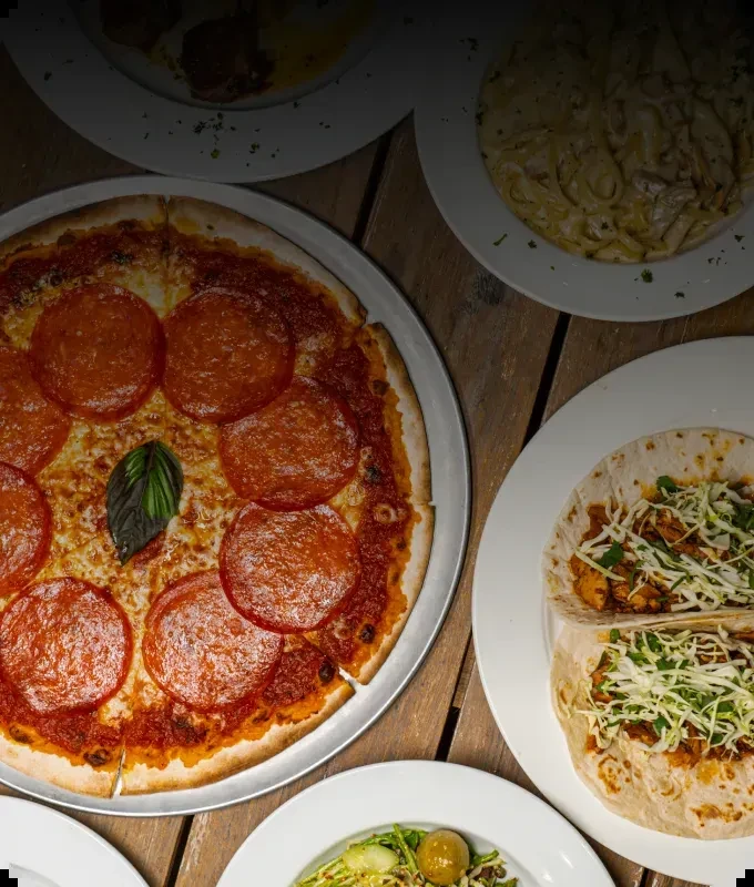 Top-down view: Pepperoni pizza, tacos, pasta, and salad on a rustic table.