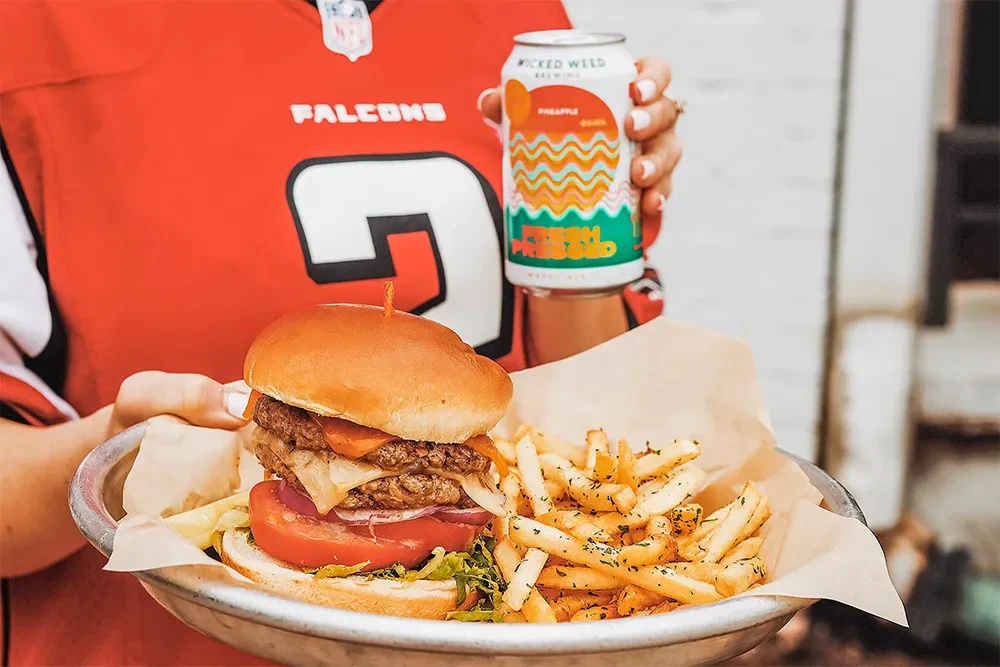 Falcons fan holding a burger, fries, and a Wicked Weed drink.