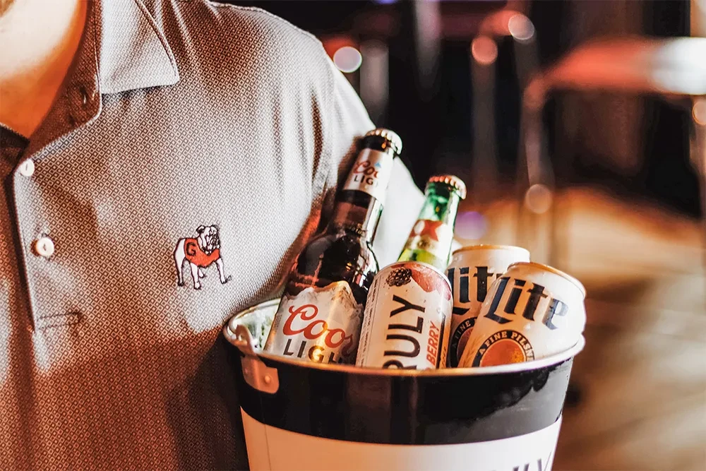 Person in grey polo, bulldog logo, holds striped bucket of drinks.