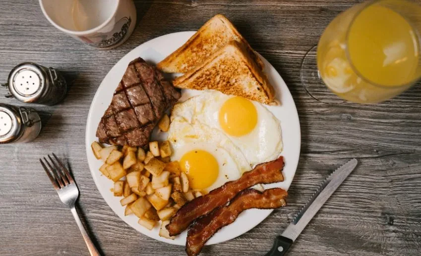 Hearty breakfast plate: steak, eggs, bacon, toast, potatoes on rustic table.