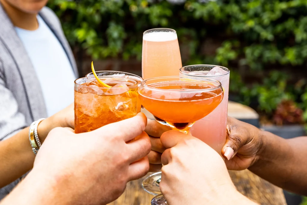 Four hands clinking various colorful cocktails outdoors.