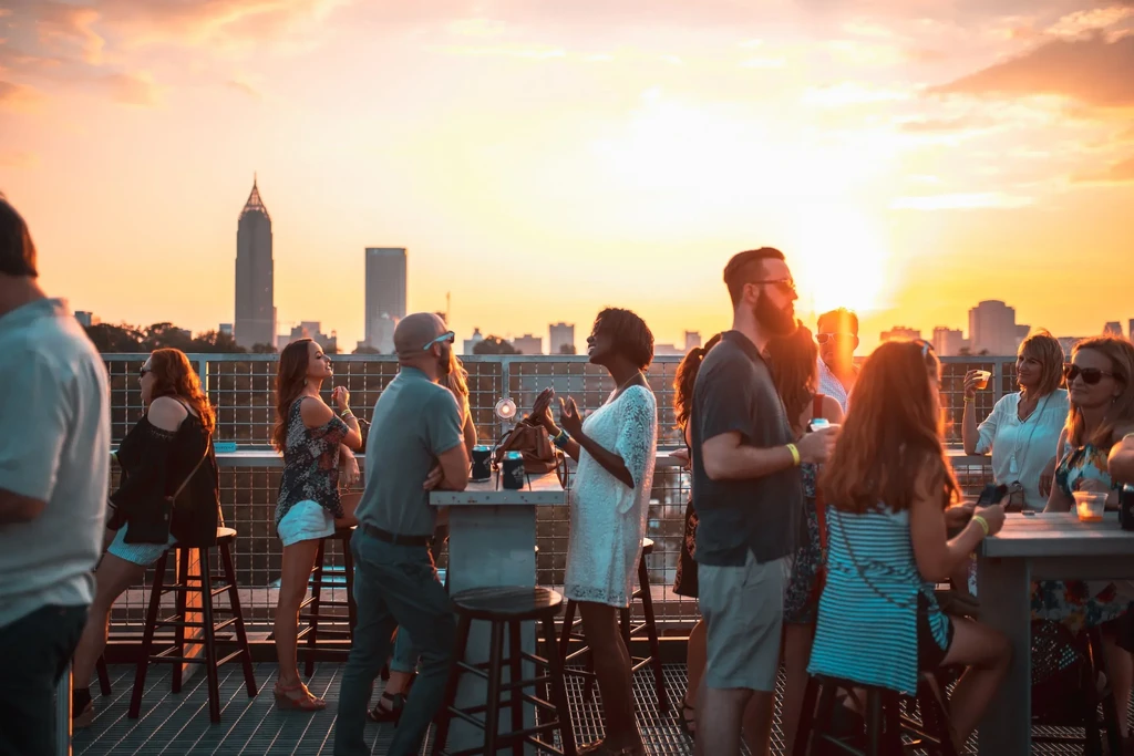 People socialize at a rooftop bar overlooking a city skyline at sunset.