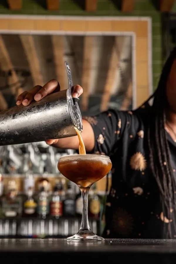 Bartender's hands pour dark amber liquid from shaker into coupe glass.