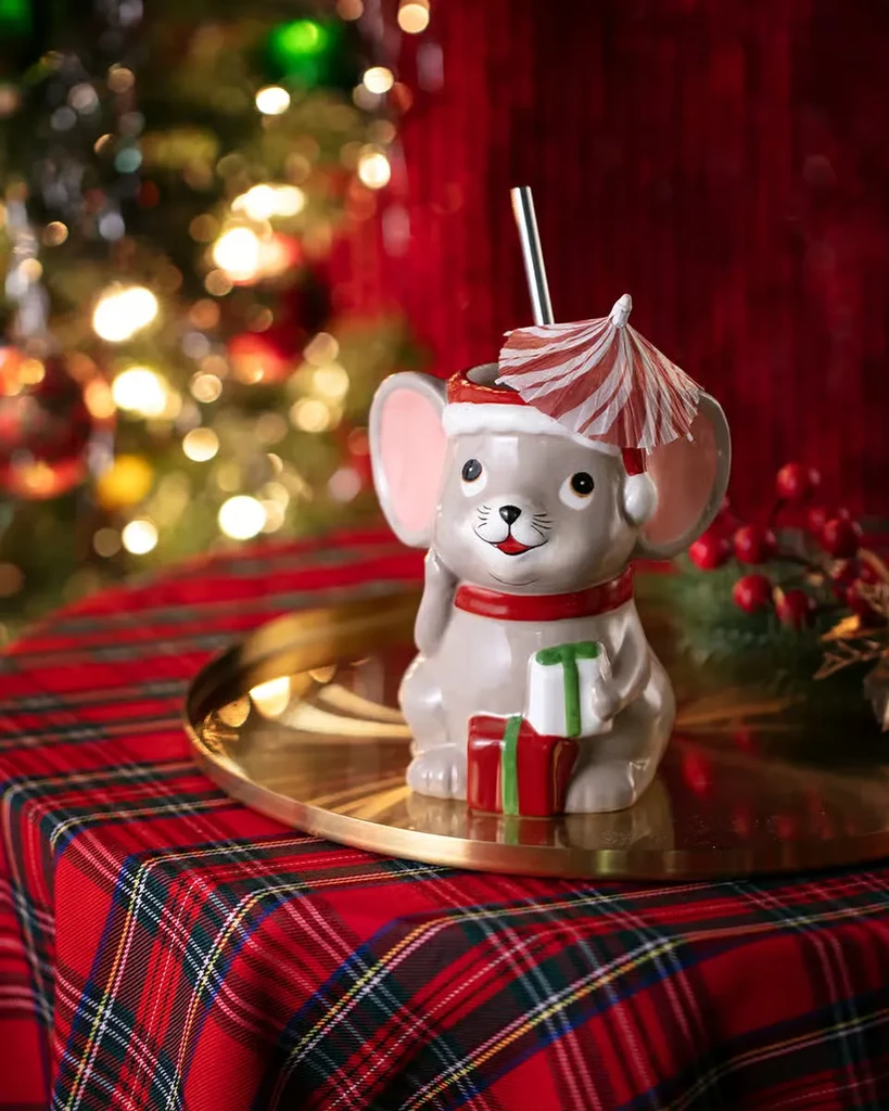 Grey mouse mug in Santa hat, drink, on plaid with blurred lights.