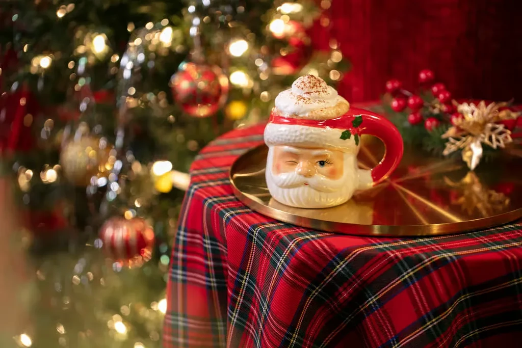 Winking Santa mug with whipped drink on plaid, festive Christmas scene.