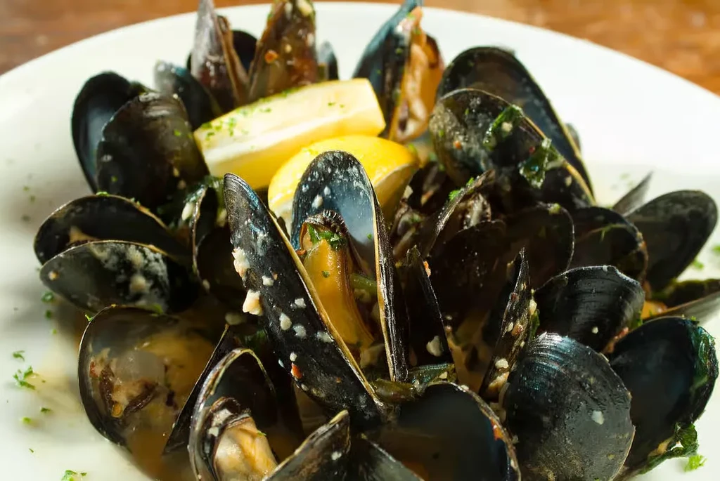 Plate of cooked mussels in sauce with herbs and lemon wedges.