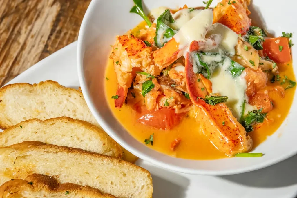 Close-up of a seafood stew with lobster, vegetables, orange sauce, and bread.