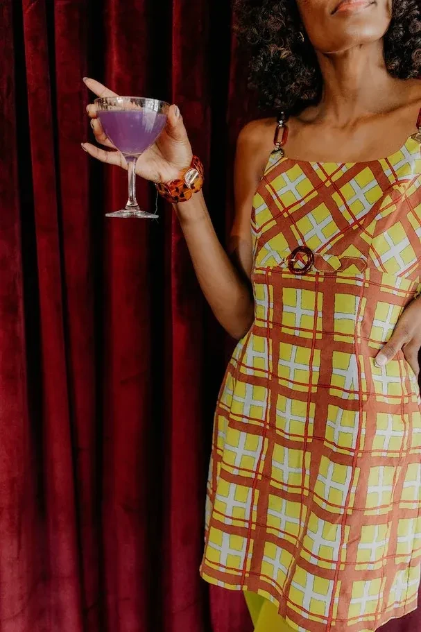Person in plaid dress holds purple cocktail by red velvet curtain.