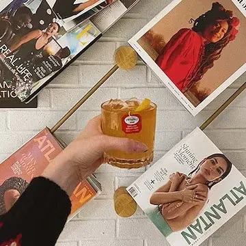 Hand holds amber iced beverage with lemon peel against a magazine wall.