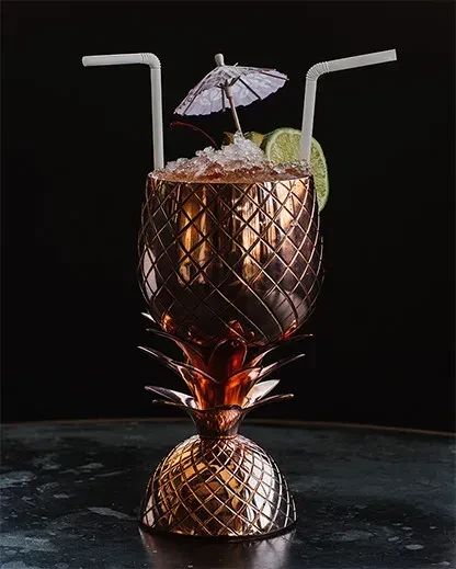 Cocktail served in metallic pineapple-shaped glass with ice and garnishes.