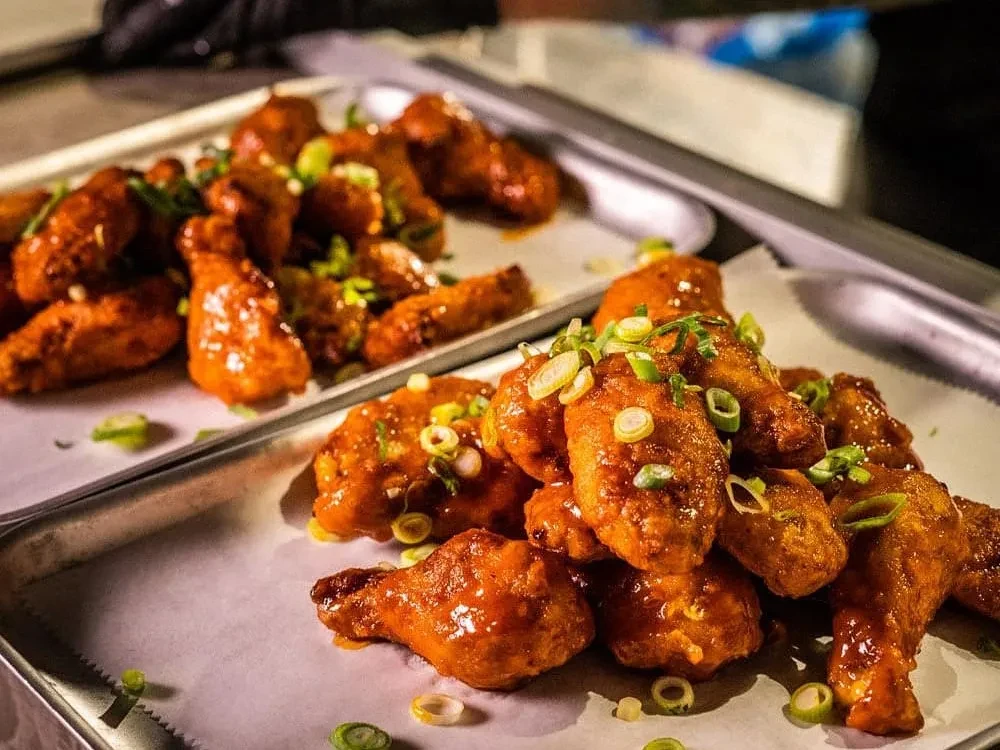 Glistening red-orange glazed chicken wings garnished with green onions on sheet pans.