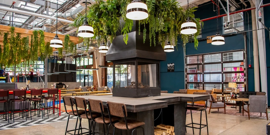 Industrial-chic restaurant interior with central fireplace, bar, and green foliage.