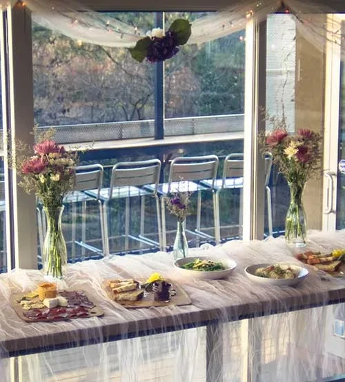 Indoor table with food and flowers, overlooking outdoor bar and bare trees.
