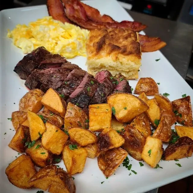Generous breakfast plate with steak, eggs, bacon, biscuit, and roasted potatoes.