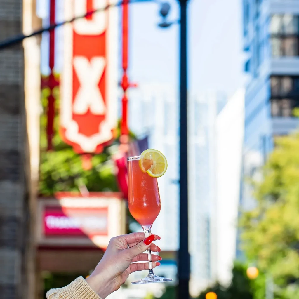 Hand holds reddish-pink cocktail with lemon slice; blurred city background.