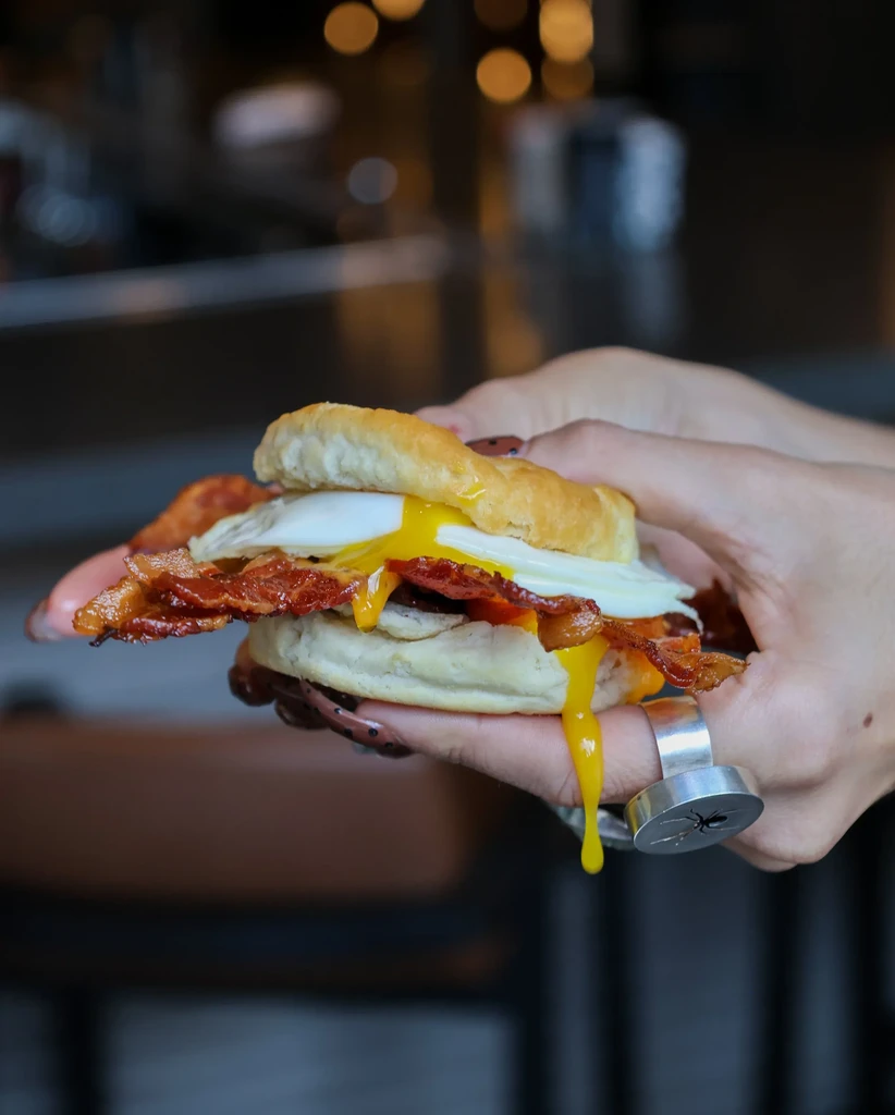 Hands hold a biscuit sandwich with crispy bacon, runny egg, and cheese.