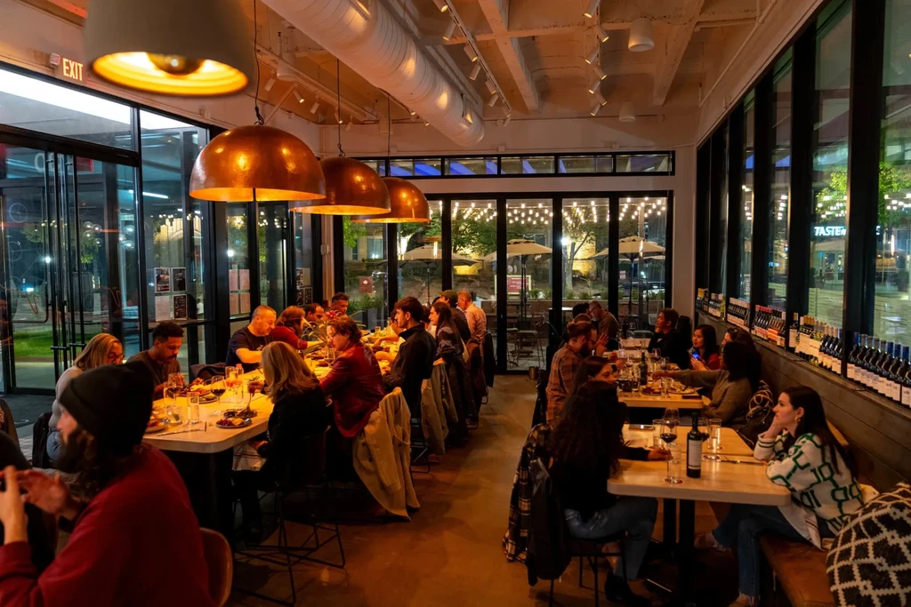 Busy contemporary restaurant interior with industrial lighting, patrons, and wine display.