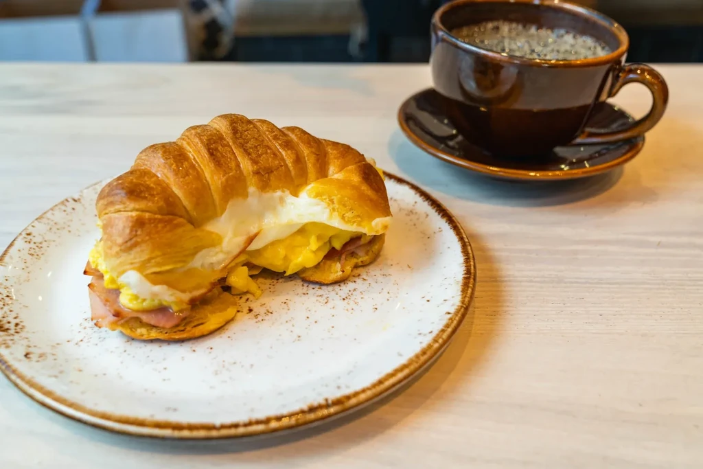 Golden-brown croissant sandwich with eggs, ham, cheese, and a coffee.