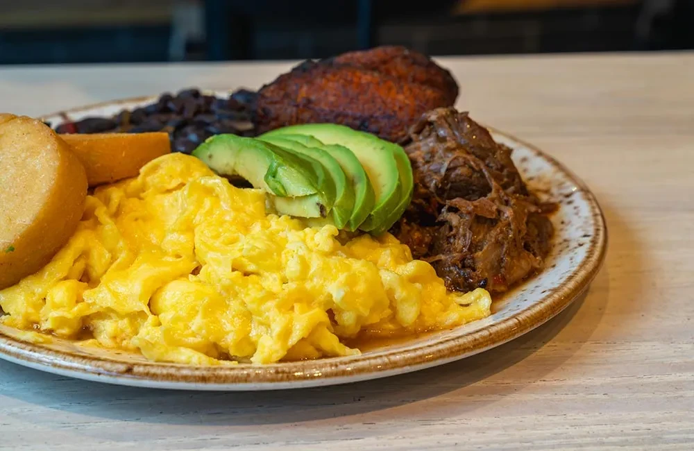 Scrambled eggs, avocado, shredded beef, beans, and plantain on a rustic plate.