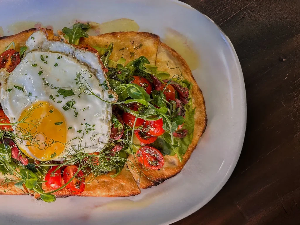 Fried egg on toasted flatbread with avocado, tomatoes, and herbs.