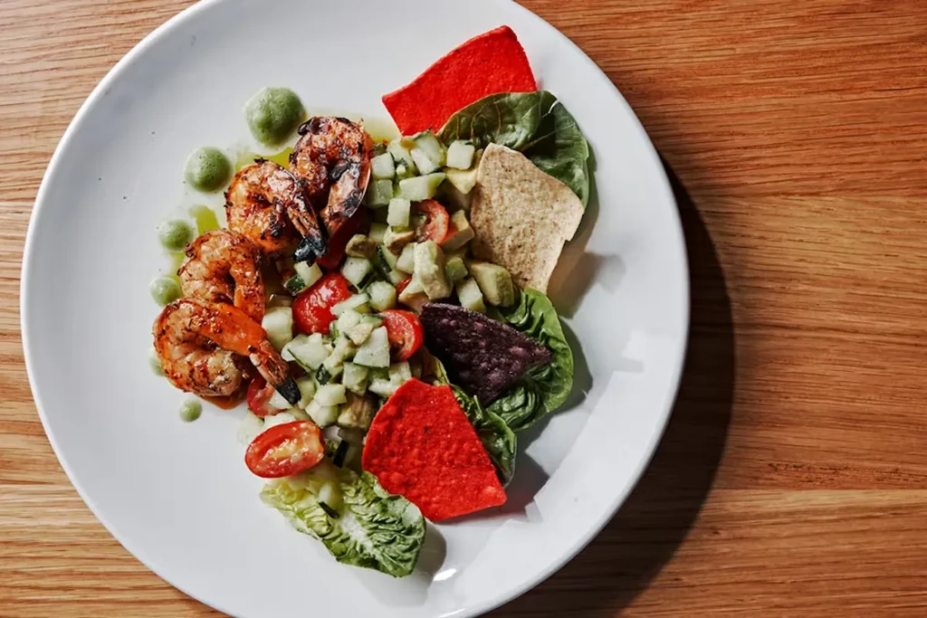 Plated dish featuring three grilled shrimp, green salsa, chips, and sauce.