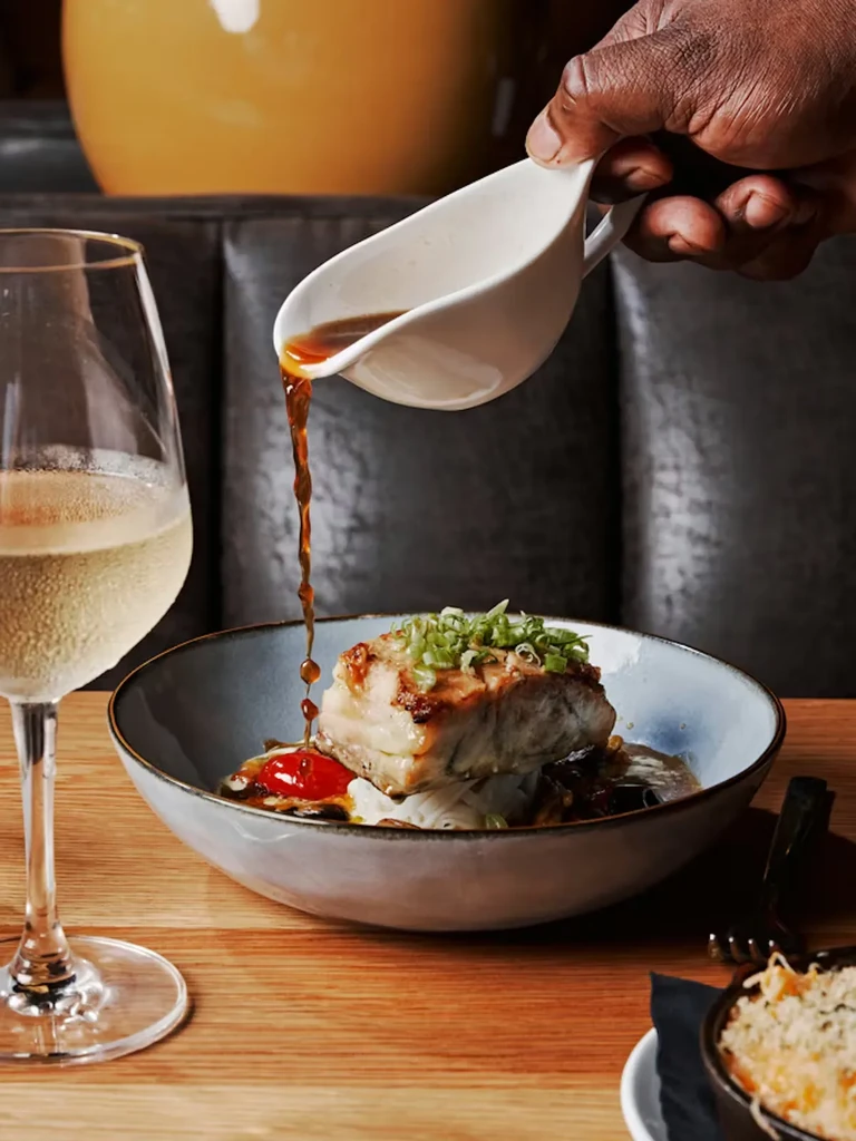 Hand pouring sauce over crispy golden dish with green onions and wine.