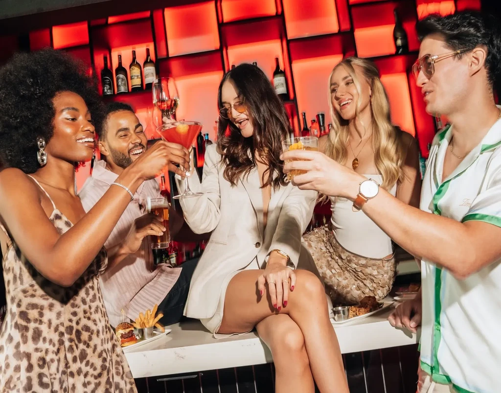 Five smiling young adults toast drinks at a bright bar.