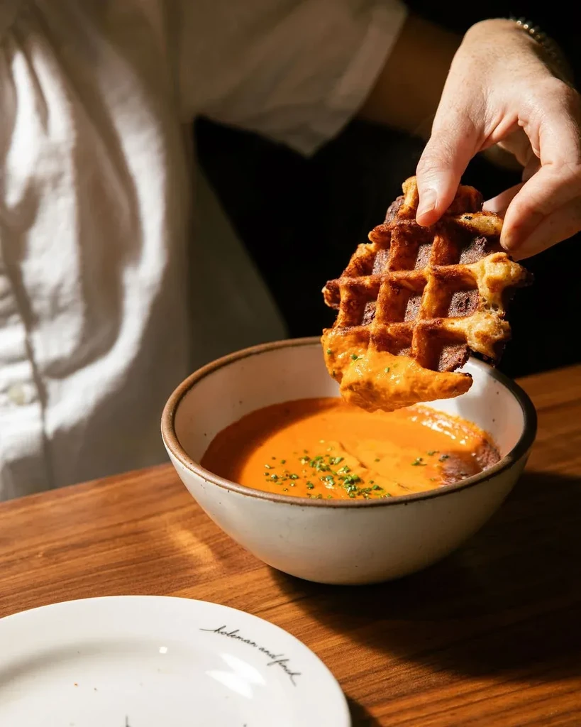 Hand dips golden waffle into creamy orange soup garnished with herbs.