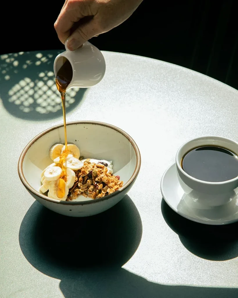 Overhead view: Hand pouring honey over yogurt, granola, bananas, and coffee.