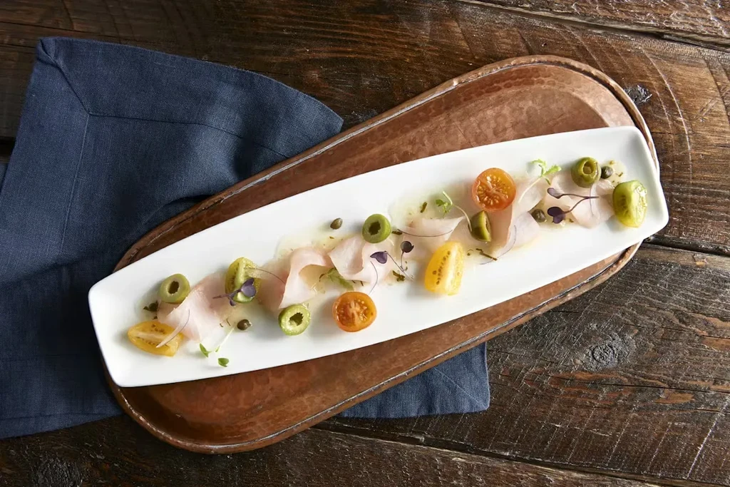 Overhead shot of seafood crudo with tomatoes, olives, capers, and microgreens.