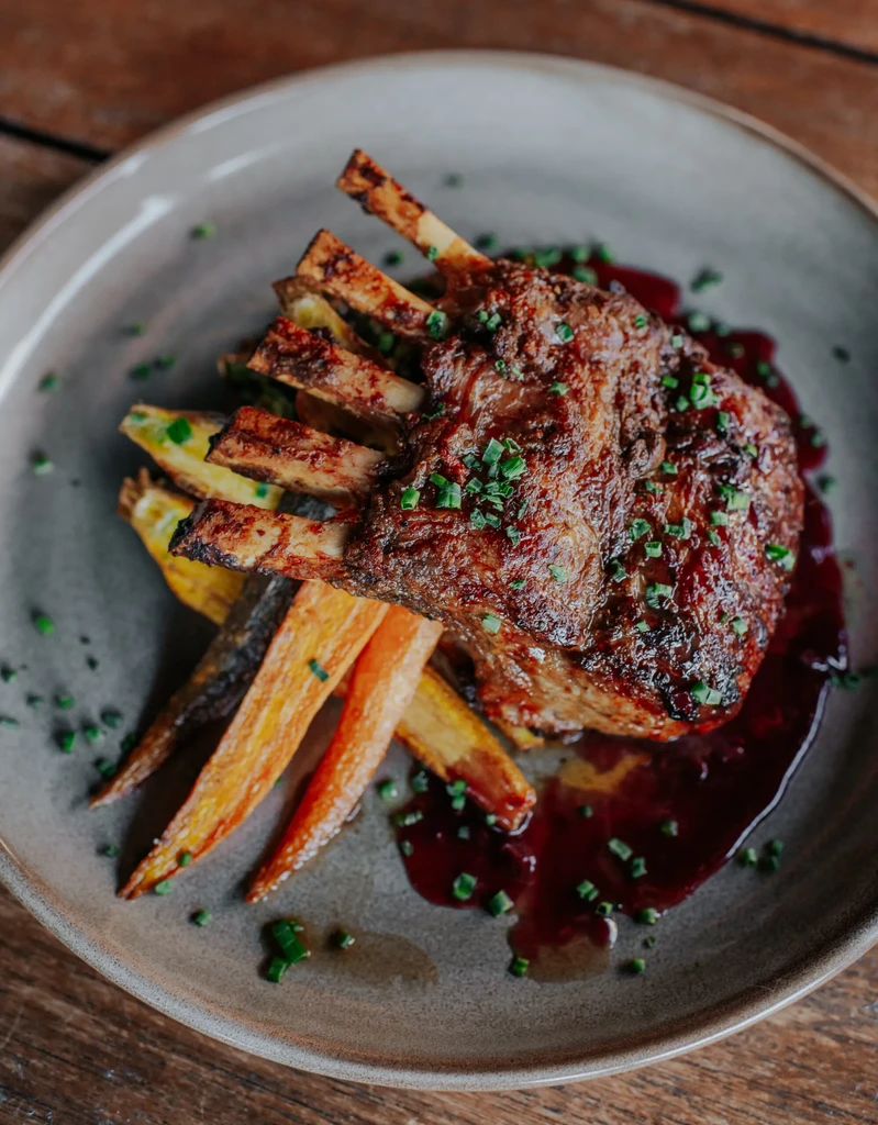 Seared rack of meat with roasted carrots, green herbs, and purple sauce.