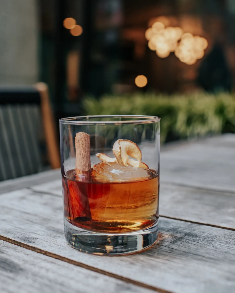 Lowball cocktail with cinnamon stick, dried fruit, ice on wooden table.