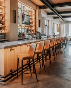 Empty modern bar with light wood counter, tan stools, and liquor display.