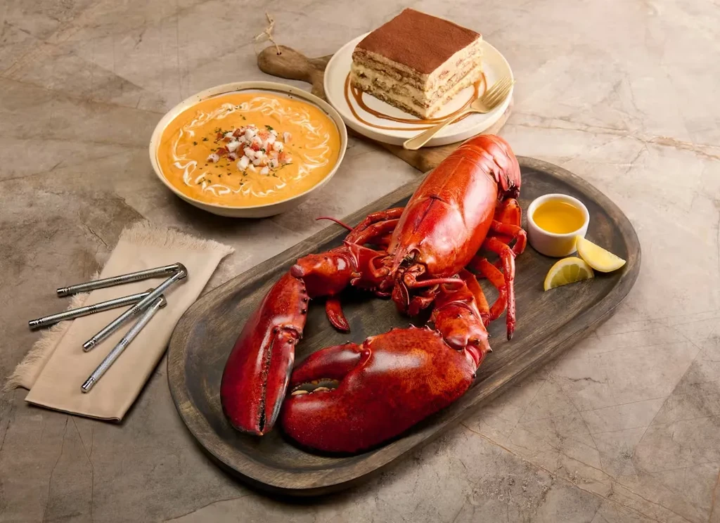 Whole cooked lobster with tools, creamy soup, and tiramisu on marble.