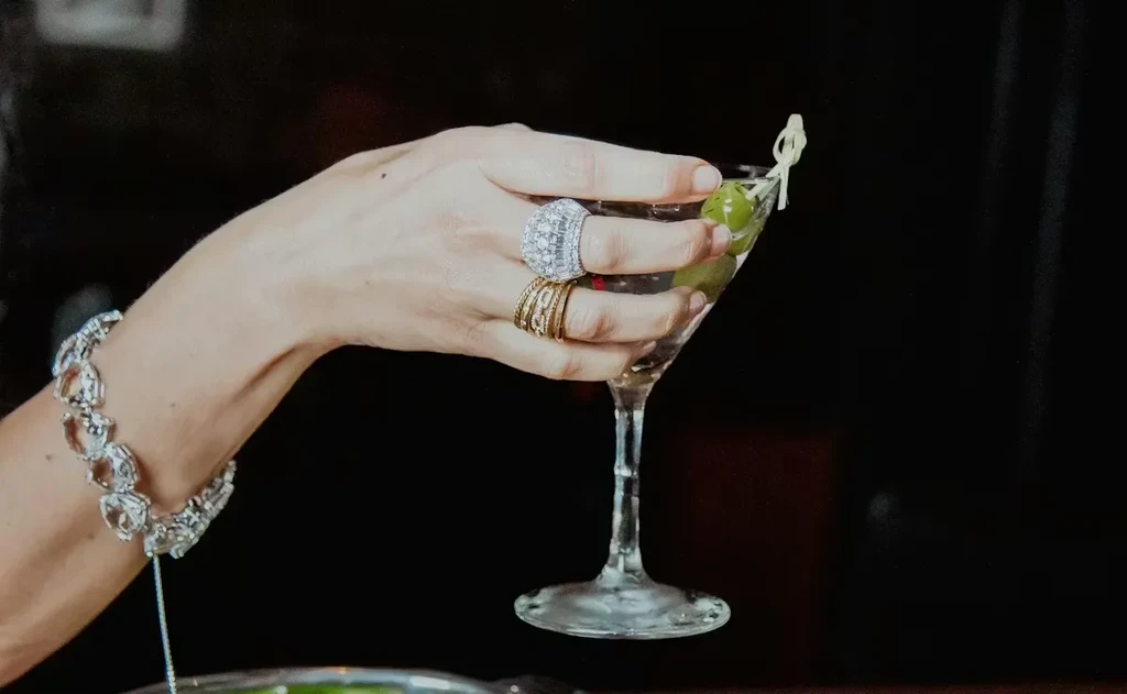 Bejeweled hand holding a martini with three olives on a dark background.