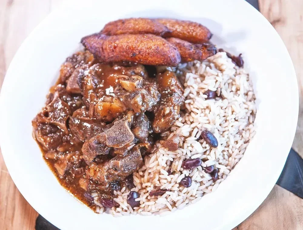 White plate with stewed meat, rice and peas, and fried plantains.
