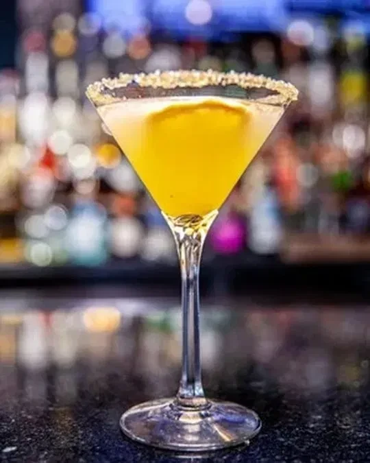 Bright yellow cocktail in martini glass with sparkling rim and citrus.