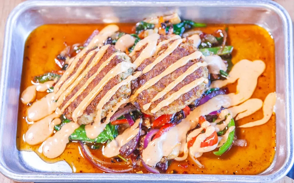 Two golden-brown patties, vegetables, and sauce in a metal tray.