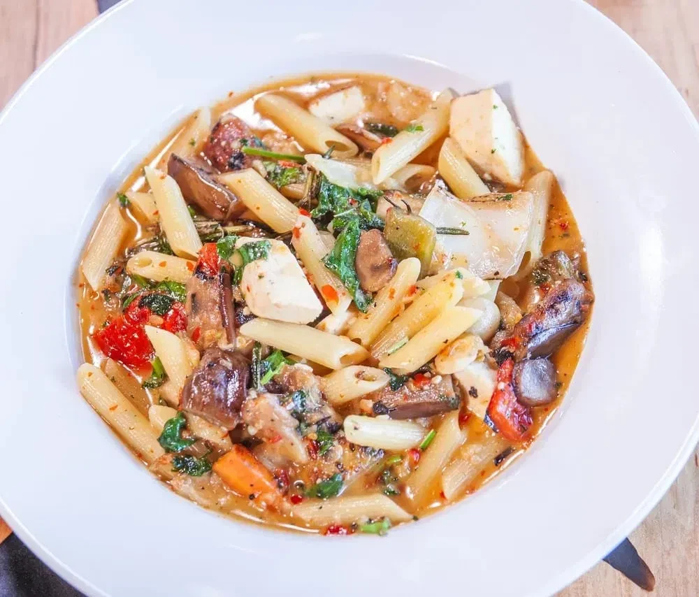 Penne pasta with mixed vegetables, white cheese, and rich reddish-brown sauce.