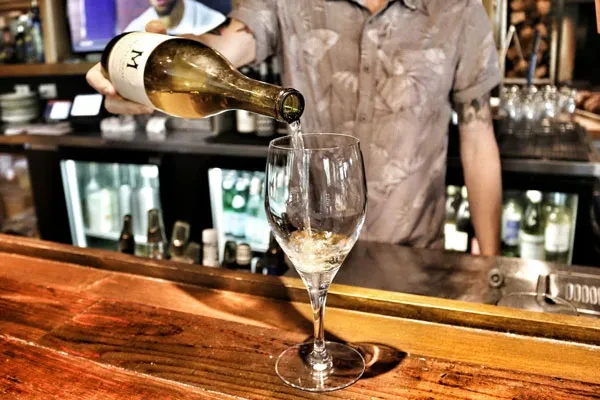 Hands pour white wine into a glass on a rustic bar.