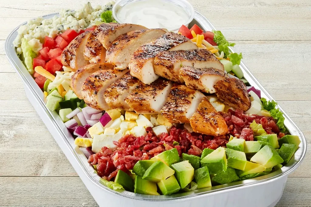 Large grilled chicken salad with many fresh toppings in a foil tray.