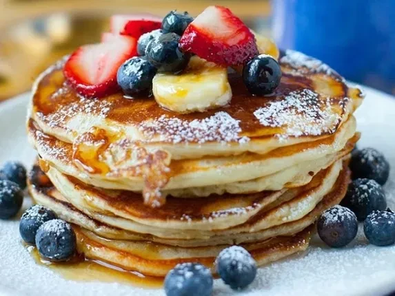 Fluffy pancake stack with syrup, strawberries, blueberries, bananas, powdered sugar.