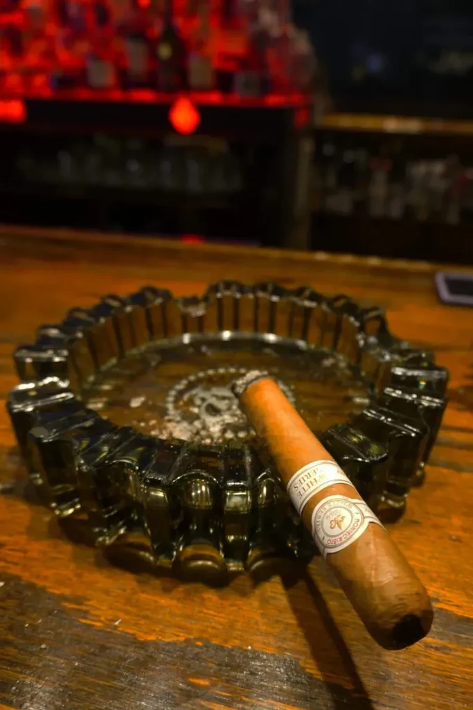 Lit Montecristo cigar in an ornate ashtray on a rustic wooden surface.