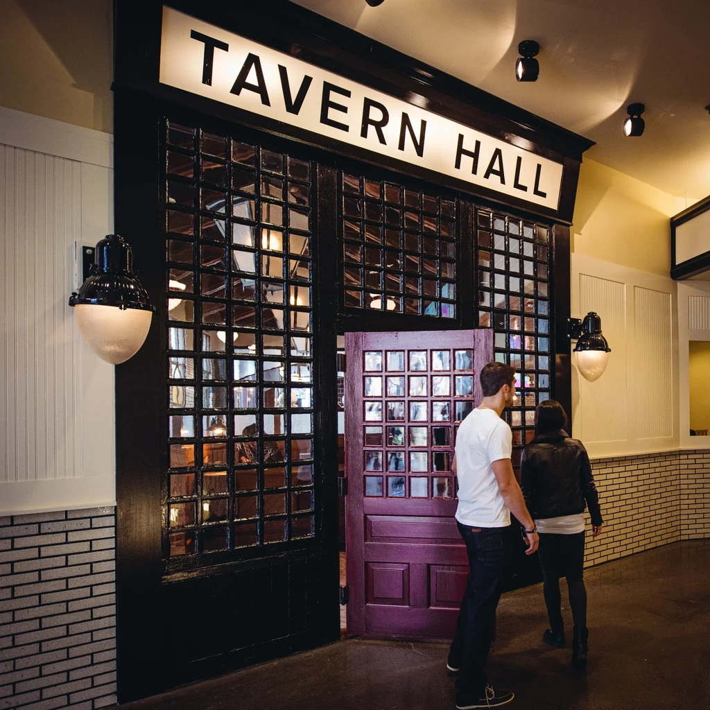 Two people enter the warmly lit TAVERN HALL restaurant entrance.