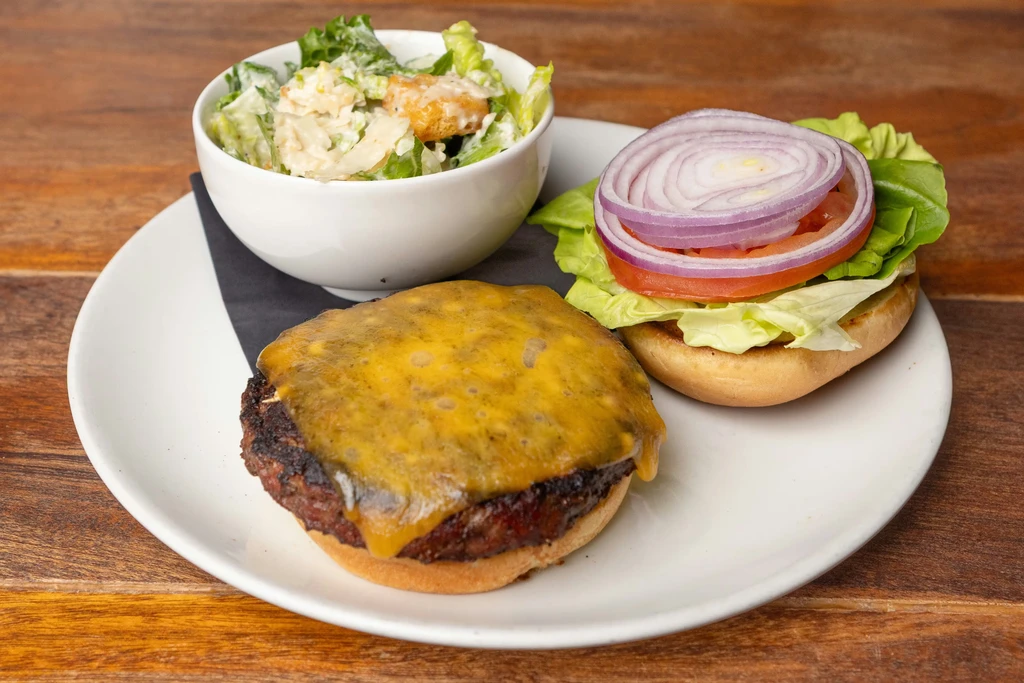 Cheeseburger with separate bun and a side Caesar salad on wood.