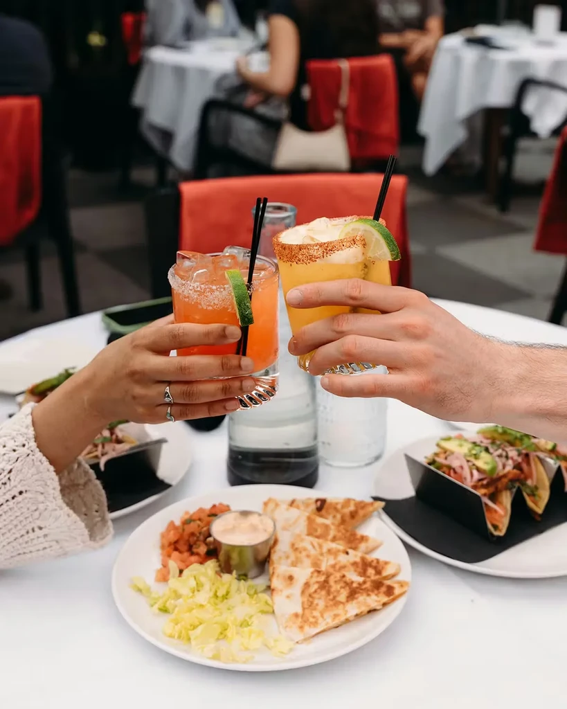 Two hands toast cocktails above quesadillas and tacos in a restaurant.