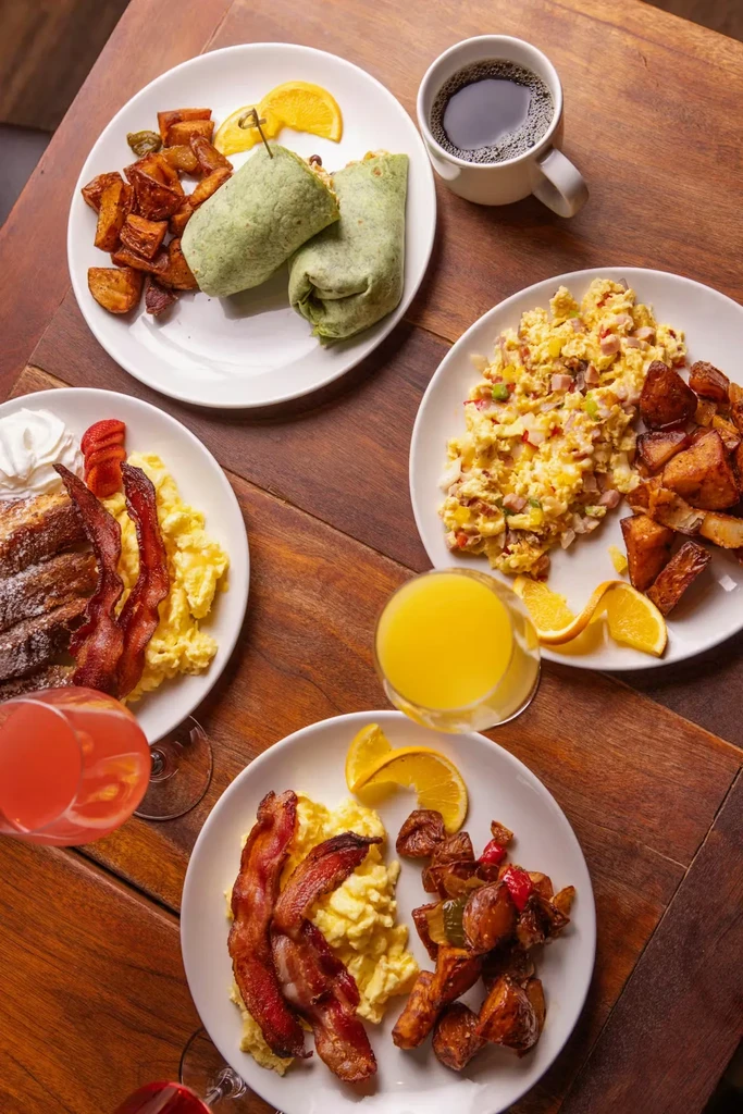 Multiple breakfast plates, coffee, juice, and mimosas on a dark wood table.