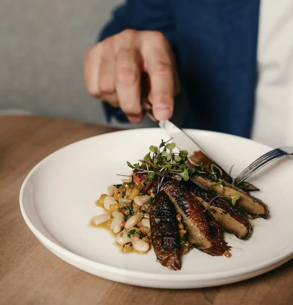 Hands cutting seared slices and white beans with microgreens on a plate.