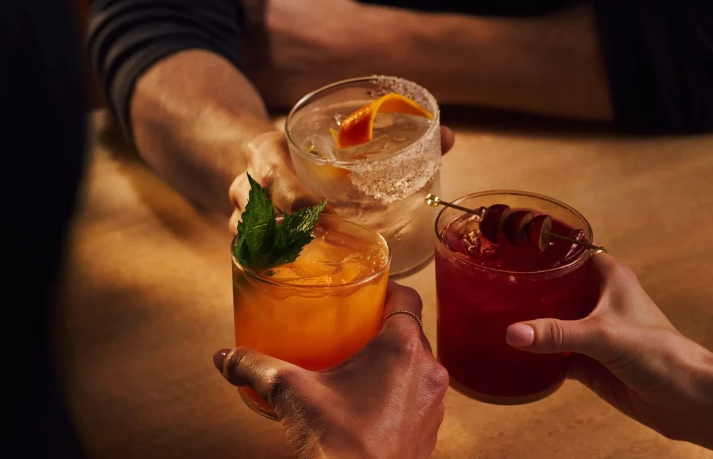 Three hands toast distinct cocktails with mint, citrus, and cherries.
